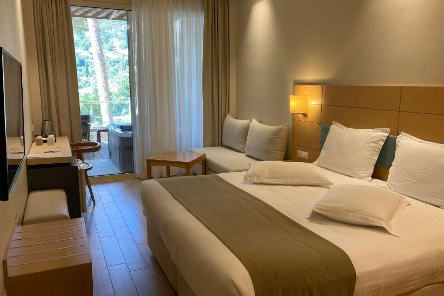 Une chambre d'amis confortable et lumineuse avec un grand lit blanc, une tête de lit en bois et un balcon donnant sur la verdure.