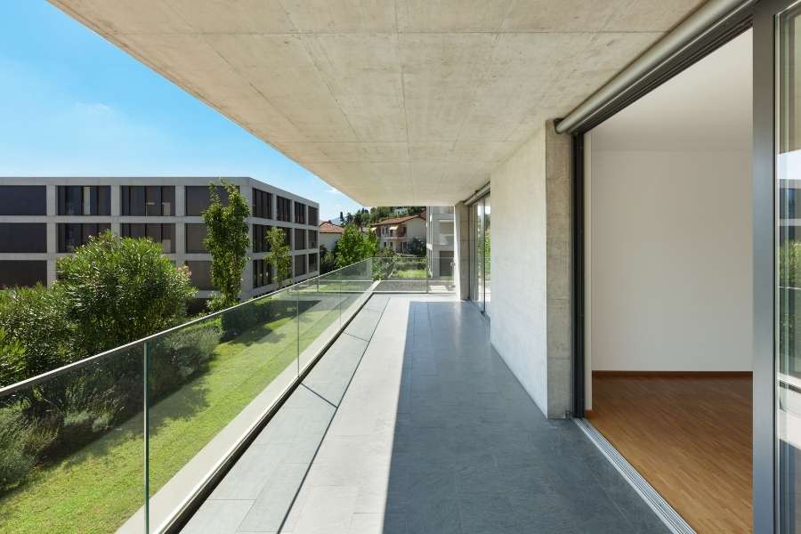 Un grand balcon moderne avec un revêtement de sol en carrelage gris uni, une balustrade en verre et une vue sur un parc.